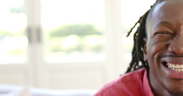 Smiling young man with braided hair wearing a red shirt, indoors with natural light. Strong positive energy, perfect for illustrating concepts of happiness, empowerment, youthful vitality, and positivity. Great for use in promotional materials, lifestyle blogs, and social media campaigns.