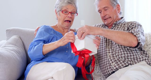 This image portrays a joyful elderly couple sitting on a couch and opening a wrapped gift with excited expressions. Suitable for use in articles or advertisements focused on senior living, family bonding, special occasions, holidays, or celebrations. Ideal for marketing relating to gifts for seniors or grandparent-related products.