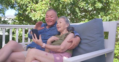 Senior asian couple enjoying video call and relaxing on porch bench with smartphone