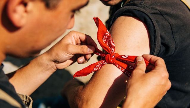 Tying red paisley bandana around upper arm, friends sharing help outdoors in warm sunlight