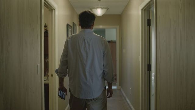 Middle-aged man walking down dim home hallway holding smartphone, back view, domestic scene
