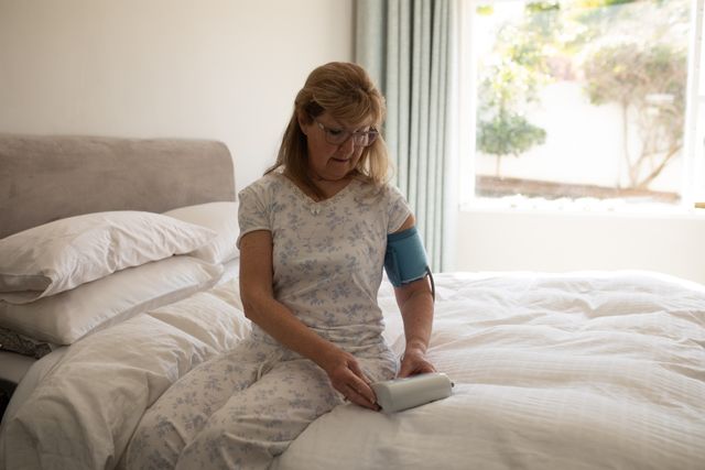 Senior Caucasian woman spending time at home, sitting in her bedroom, checking her blood pressure in the bed. Social distancing during Covid 19 Coronavirus quarantine lockdown.