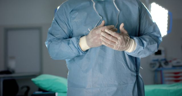 Medical professional wearing blue scrubs and sterile gloves, standing in an operating room, hands clasped together. Ideal for use in healthcare, medical procedures, surgery preparation, and hospital-related themes.