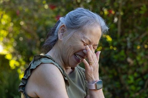 Joyful senior asian woman laughing while covering face in sunlit garden with soft bokeh