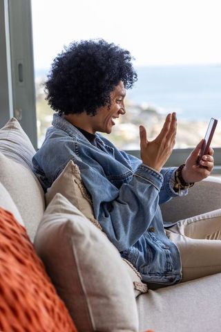 Smiling african american man video calling on smartphone while relaxing by sea-view window