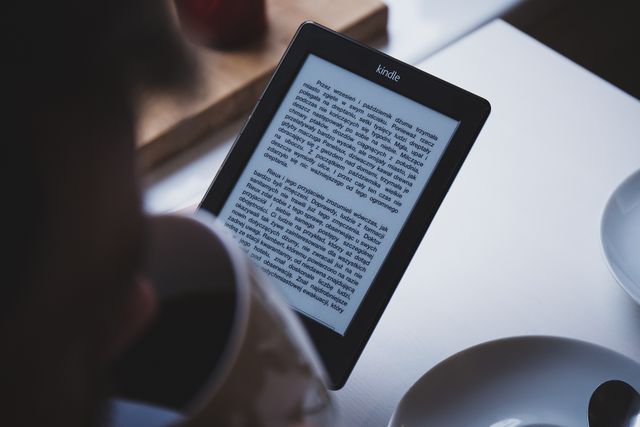 A man is reading a digital book on a Kindle while holding a coffee mug. The setting appears relaxed and comfortable, suitable for enjoying a peaceful reading experience. This image is ideal for promoting e-reading devices, digital literacy, coffee shops, cozy indoor environments, and relaxation themes.