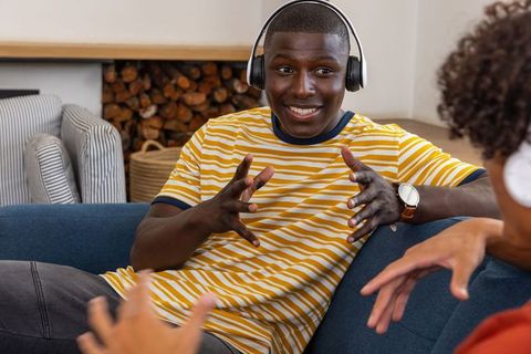 African American friends wearing headphones chatting on cozy living room sofa