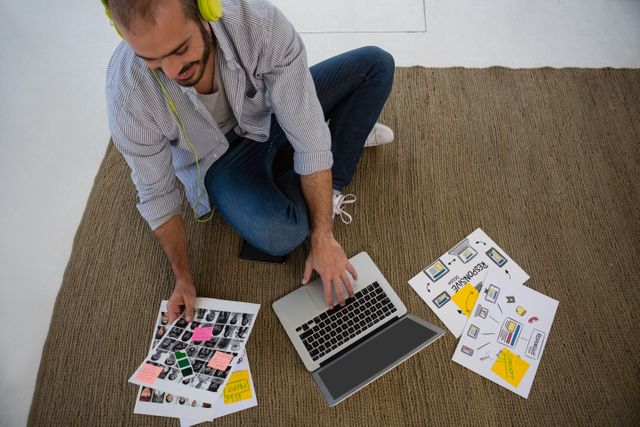 Designer sitting on floor using laptop with creative materials spread around. Ideal for illustrating creative workspaces, design processes, brainstorming sessions, and modern work environments. Useful for articles, blogs, and advertisements related to design, creativity, and remote work.