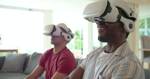 Diverse friends enjoying VR gaming, smiling on couch at home with white headsets, sunlight