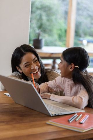 Mother and daughter smiling while studying on laptop, warm home tutoring and bonding