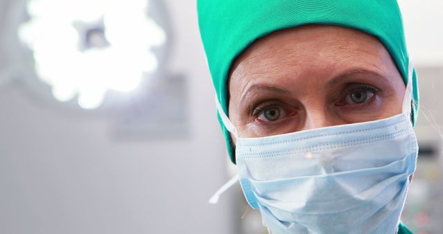 This image shows a close-up view of a surgeon wearing a surgical mask and cap, looking focused while in an operating room. This can be used in medical publications, hospital websites, healthcare promotions, articles about surgery, and educational materials for medical students.