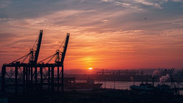 Sunset silhouetting gantry cranes over industrial port with container ships and orange sky