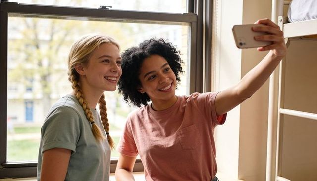 Sunlit college roommates taking selfie in cozy dorm room smiling teens sharing friendship
