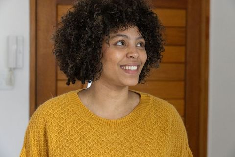 Smiling young woman wearing mustard-yellow sweater looking toward entryway with intercom