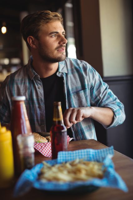 Young man in plaid shirt enjoying a meal at a restaurant. He is eating a burger and fries, with condiments like ketchup and mustard on the table. Ideal for use in advertisements for restaurants, food blogs, or lifestyle articles about dining out.