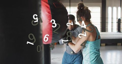 Female trainee punching heavy bag while male coach holds red-black pad in boxing gym ring
