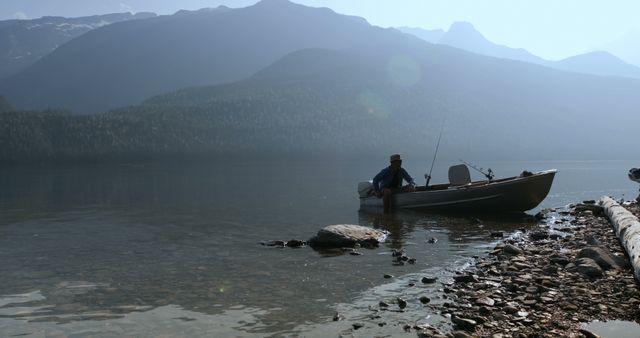 Person sitting in boat near lake shore surrounded by misty mountains and serene water. Great for outdoor, adventure, and nature-themed content. Ideal for promoting summer vacations, recreational fishing, and relaxation.
