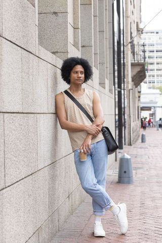 African american man leaning on stone wall holding coffee and black bag on downtown sidewalk