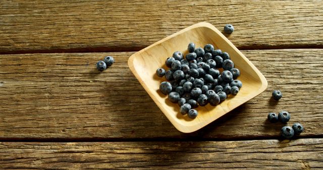 A square wooden bowl filled with fresh blueberries sits on a rustic wooden table, with copy space. Blueberries are known for their health benefits and are often included in discussions about superfoods.