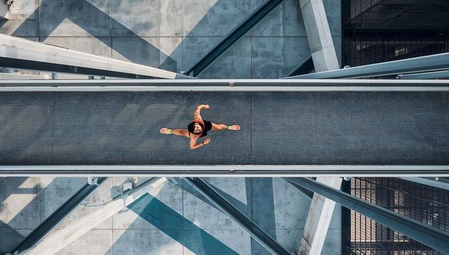 Overhead urban runner crossing metal skywalk in neon shoes with geometric shadows