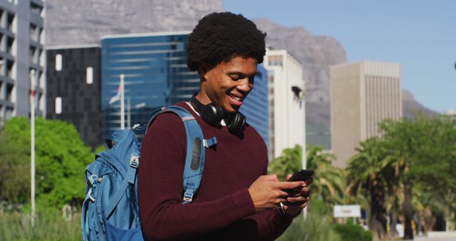 Smiling african american man using smartphone, wearing headphones and backpack in city street. digital nomad on the go, out and about in the city.