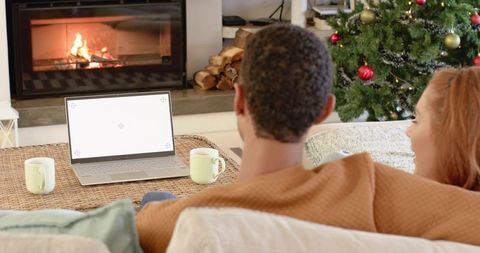 Diverse couple relaxing by fireplace, watching laptop with mugs and christmas tree