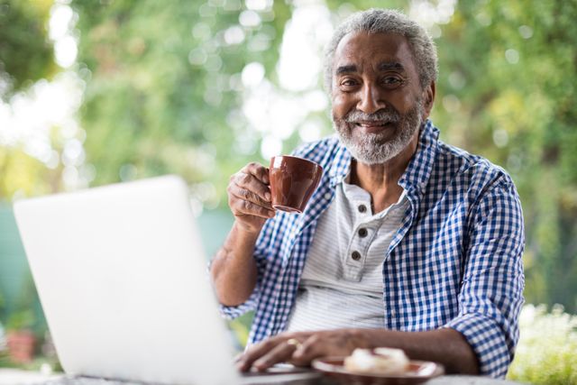 Senior man sitting at a table in a garden, holding a coffee cup and using a laptop. He is smiling and appears relaxed, enjoying his morning routine. This image can be used for promoting senior lifestyle, technology use among elderly, or leisure activities.