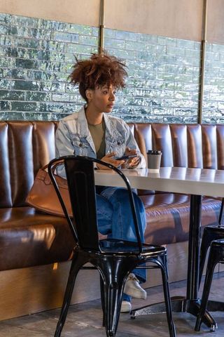 African american woman sitting in cafe booth using smartphone, holding coffee, denim jacket