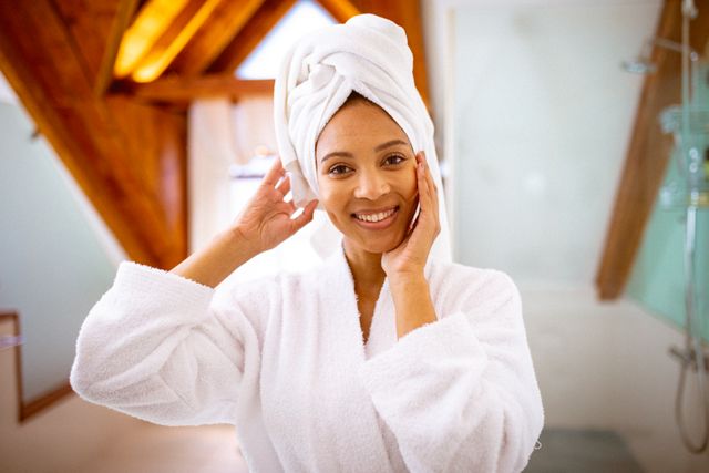 This image depicts a smiling woman wearing a bathrobe and towel turban, enjoying a moment of self-care at home. Ideal for use in articles or advertisements related to skincare, wellness, beauty routines, and domestic lifestyle. Perfect for promoting self-care products, morning routines, and relaxation techniques.