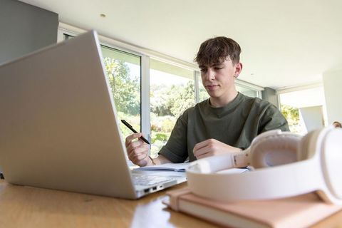Teen studying at home with laptop and headphones taking notes by window in natural light