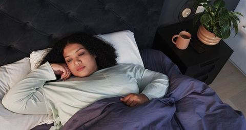 Mid-adult woman resting in mint sleep top under purple duvet in modern cozy bedroom