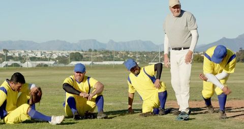 Male baseball team resting with veteran coach on sunny suburban outfield