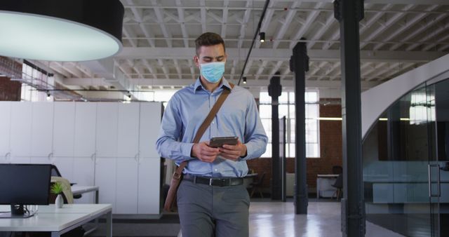 Ideal for illustrating safe business practices during the COVID-19 pandemic, this image shows a professional young adult man walking through a modern office while checking his smartphone. Perfect for articles or content on business continuity, safety protocols, or remote work integration.