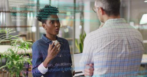 Woman explaining code to colleague in modern office holding laptop with code overlay