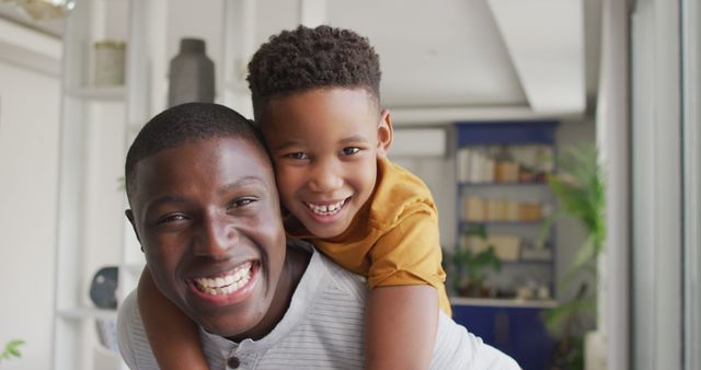 This image shows an African American father and son smiling and enjoying each other's company indoors. The father and son appear to be very happy, showcasing the strong bond between them. The background features a modern, well-lit living space. This is perfect for use in advertisements and promotions about family life, parenting, family bonding, and positive emotions.