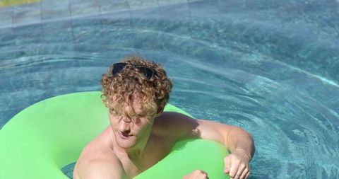 Curly-haired shirtless man floating on neon green inflatable tube in backyard pool on sunny day