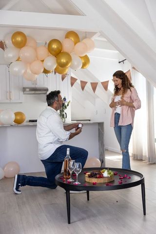 Man kneeling proposing, woman reacting in bright loft kitchen with balloons and wine