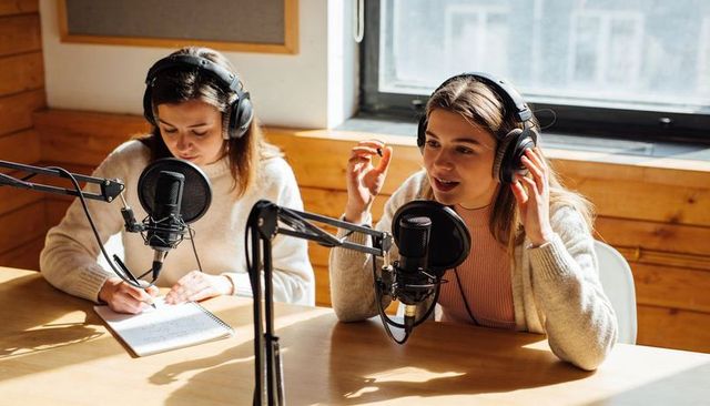 Recording two co-hosts conversing in sunlit acoustic podcast studio wearing headphones, taking notes