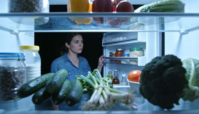 Woman peering into fridge at night holding door, searching for food, moody blue light