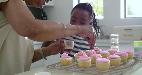 Grandmother and granddaughter decorating pink frosted cupcakes together in home kitchen