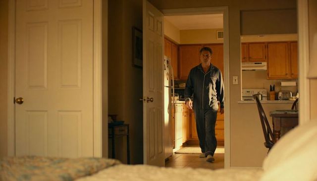 Middle-aged man walking from small kitchen into bedroom wearing dark pajama set in cozy home