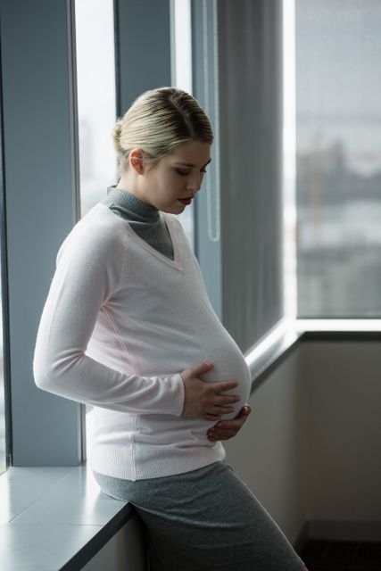 Pregnant businesswoman standing by window in modern office, touching her belly and looking thoughtful. Ideal for use in articles about maternity leave, balancing work and pregnancy, corporate wellness programs, and women's health in the workplace.