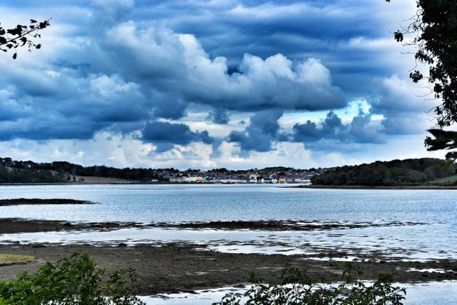 Dramatic sky with dense clouds reflecting in the calm sea water near the shoreline. Captures a scenic coastal area with forested land in the background offering a peaceful and moody atmosphere. Perfect for depicting nature's beauty, travel destinations, and tranquil scenery.