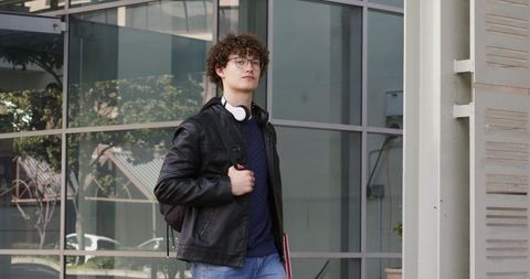 Curly-haired student walking with backpack and red binder near modern glass facade