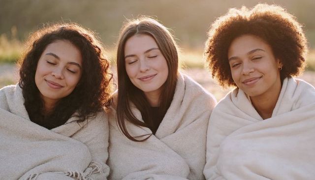 Three women snuggling in beige blankets smiling with closed eyes at golden hour in field