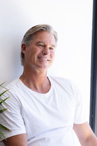 Serene mature man smiling by bright window wearing white t-shirt, relaxed senior portrait