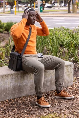 Young african american man adjusting headphones while sitting on concrete bench in urban park listen