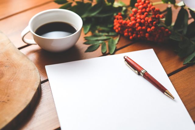 Blank sheet of paper with red pen and coffee cup on wooden table accompanied by red berries. Use for concepts like writing, morning routine, new beginnings, and creative workspace.