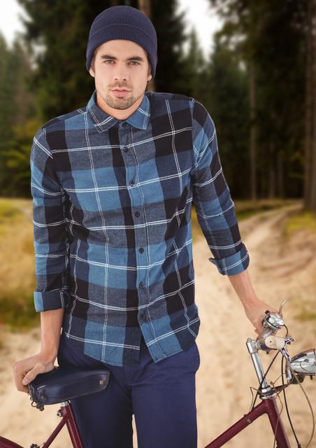 The man is standing on a forest pathway, holding a bicycle. He is wearing a blue plaid shirt and a beanie, presenting a casual and stylish look perfect for an outdoor adventure. This image can be used for promoting outdoor activities, travel destinations, lifestyle blogs, fashion brands targeting young adults, or health and wellness campaigns encouraging active living.