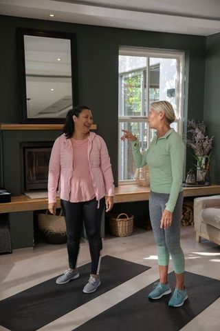 Senior women chatting during home workout on yoga mats by fireplace in cozy living room